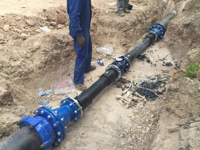 Excavadora amarilla cavando una zanja en un camino de tierra, con árboles y colinas al fondo.