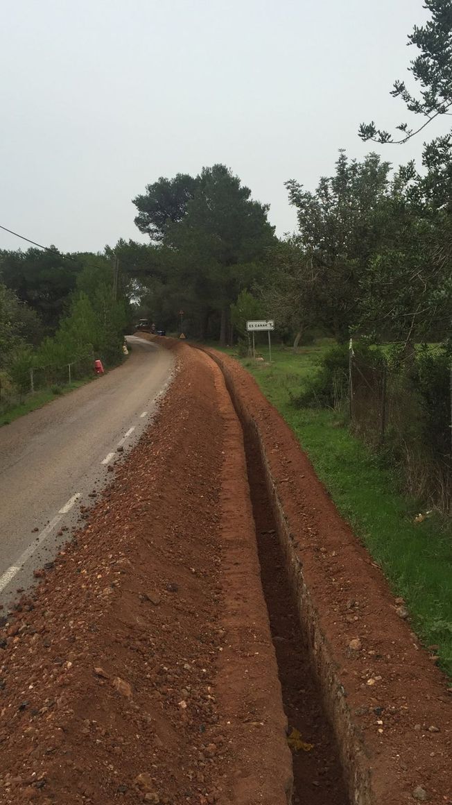Un camino con una zanja profunda cavada a su lado, con árboles y un cielo nublado al fondo.