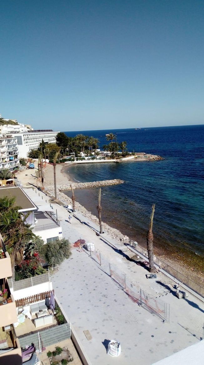 Vista de una ciudad costera con edificios blancos, una playa y agua azul clara bajo un cielo brillante.