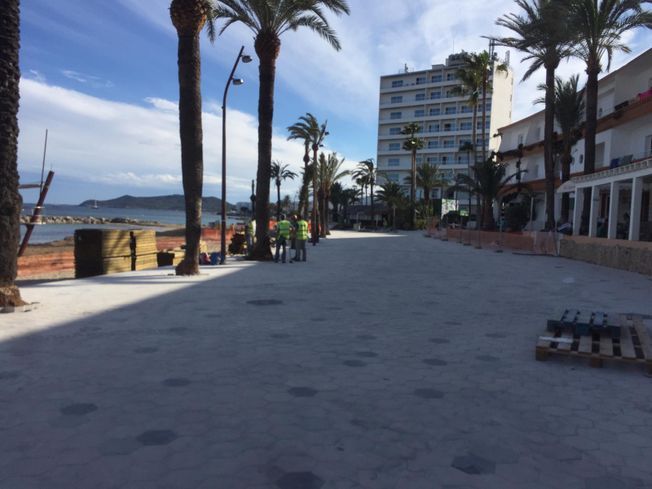 Un amplio paseo marítimo gris en construcción, con palmeras y un edificio a lo lejos.