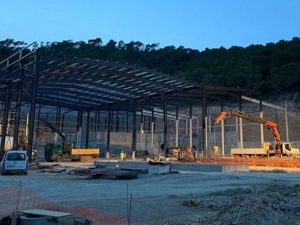 Estructura de acero de un gran edificio en construcción con maquinaria y árboles al fondo.