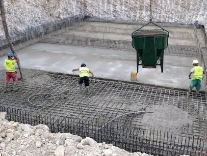 Obreros de la construcción vierten hormigón en una base, utilizando un contenedor suspendido; barras de refuerzo visibles.