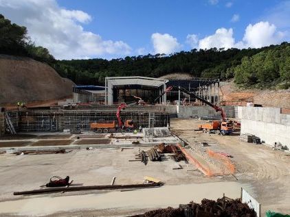 Sitio de construcción con grúas, camiones y estructuras parcialmente construidas; fondo de ladera bajo un cielo nublado.