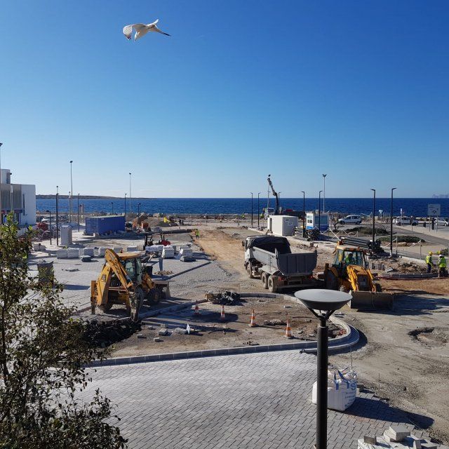 Sitio de construcción al amanecer; excavadora, trabajadores y edificio al fondo.