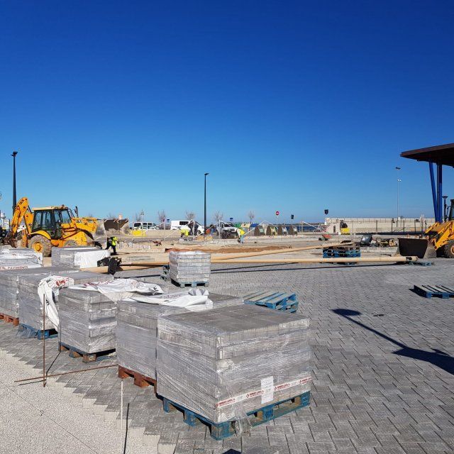 Sitio de construcción con pilas de adoquines grises, maquinaria amarilla y un cielo azul.
