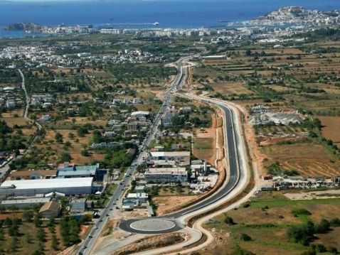 Vista aérea de una ciudad costera con una carretera de varios carriles que serpentea a través de campos; edificios y el mar en la distancia.