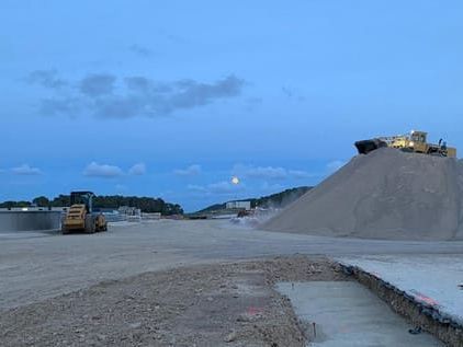 Sitio de construcción al anochecer con maquinaria pesada, incluido un cargador sobre un montón de arena bajo un cielo azul con la luna.