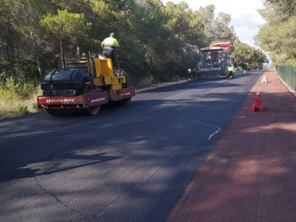 Camino en proceso de repavimentación, bordeado de árboles y con un muro bajo de piedra, conos de construcción a la vista.