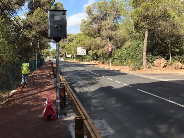 Cámara de velocidad junto a un carril bici. Un ciclista pasa. Árboles y señales de tráfico al fondo.