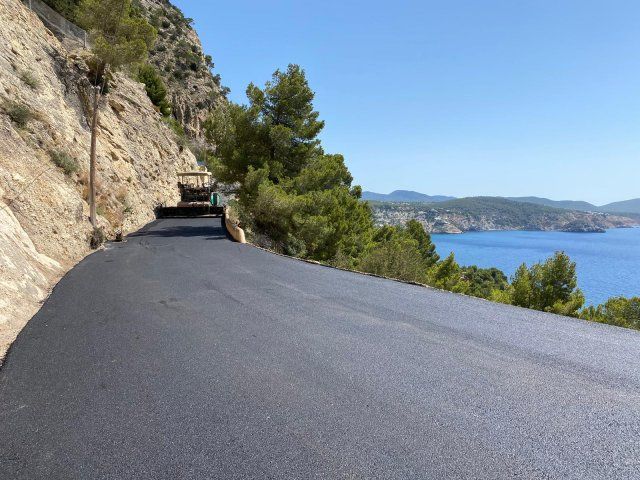 Pavimentadora asfáltica colocando una carretera costera. Cielo azul, mar y montañas de fondo.
