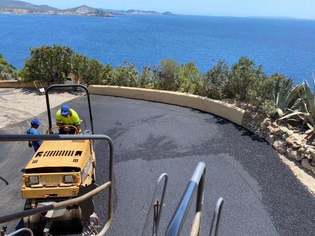 Obreros pavimentan una carretera con curvas con el mar al fondo; un rodillo amarillo compacta asfalto fresco.