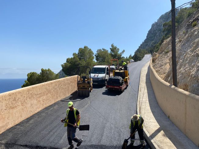 Construcción de carreteras: Obreros pavimentando una sinuosa carretera costera. Dos apisonadoras y una furgoneta a lo lejos, bajo un cielo azul.