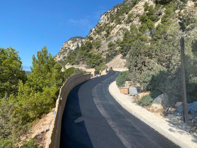 La carretera asfaltada serpentea por una ladera con árboles y un cielo azul.