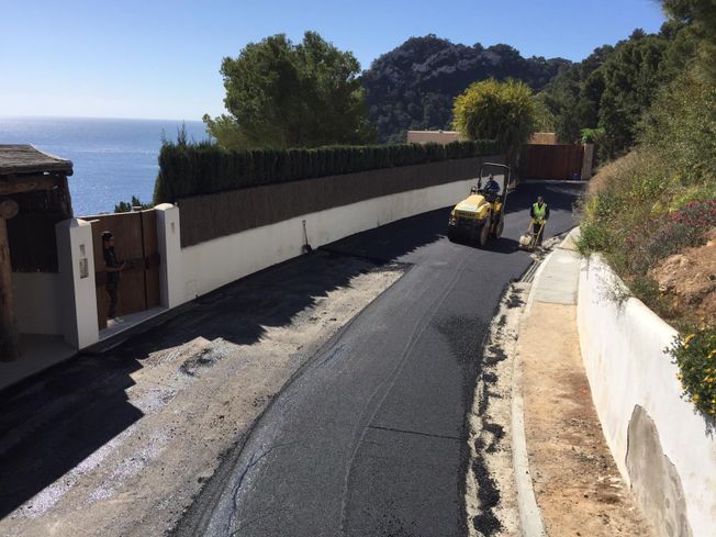 Pavimentación de una carretera asfaltada cerca de la costa con trabajadores, un rodillo y vistas al mar y las colinas.