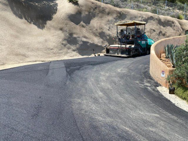 Máquina pavimentadora de asfalto colocando una nueva superficie de carretera en una ladera con tierra color canela y edificio.