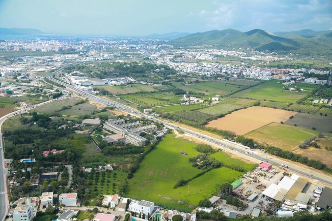 Vista aérea de una ciudad con carreteras, edificios y campos verdes, montañas al fondo bajo un cielo nublado.