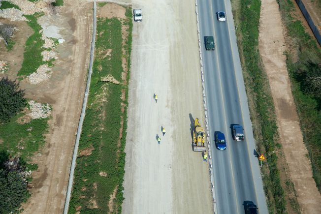 Vista aérea de la construcción de una carretera. Una excavadora amarilla y trabajadores recorren un camino de tierra junto a una carretera asfaltada con vehículos.