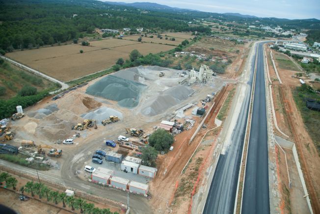 Sitio de construcción de una carretera con montones de grava, maquinaria pesada y edificios temporales al lado de una carretera recién pavimentada.