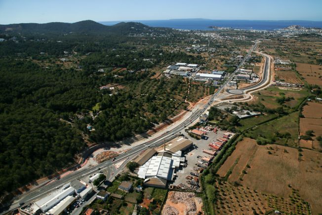 Vista aérea de una carretera que atraviesa verdes colinas y campos, con edificios a lo largo de la carretera y el océano visible.