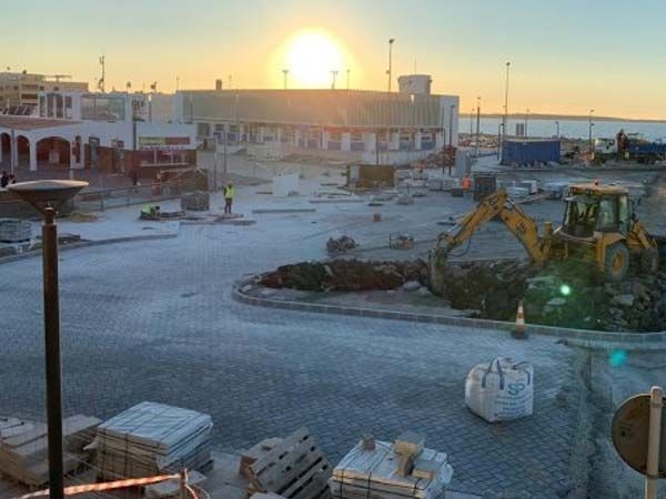 Sitio de construcción al amanecer; excavadora, trabajadores y edificio al fondo.