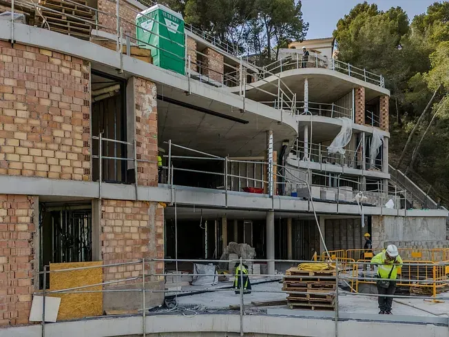 Obra de construcción de un edificio de varias plantas con elementos de ladrillo y hormigón. Hay trabajadores con chalecos de seguridad.