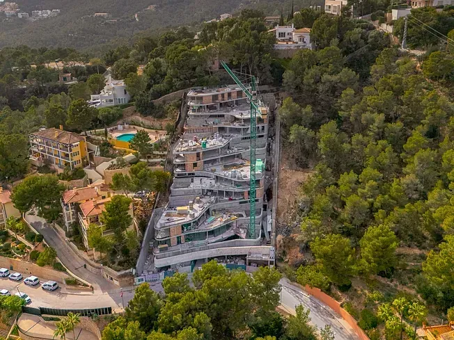 Sitio de construcción con edificios, grúa en una ladera con árboles verdes y casas circundantes.