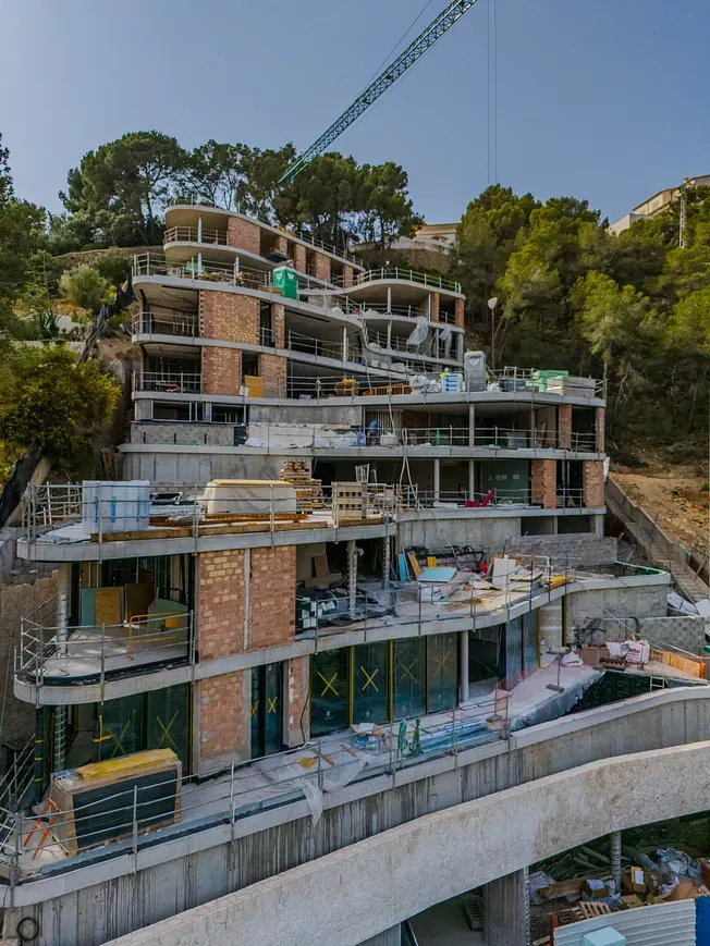Construcción de un edificio de varios pisos en una ladera, con grúa, hormigón y ladrillos visibles.