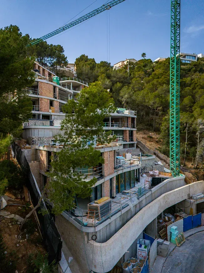 Sitio de construcción de un edificio de varios pisos en una ladera, con una grúa verde encima.