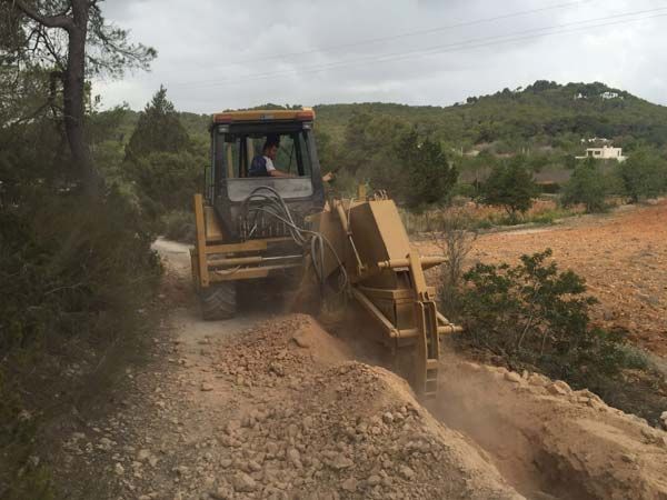 Excavadora amarilla cavando una zanja en un camino de tierra, con árboles y colinas al fondo.