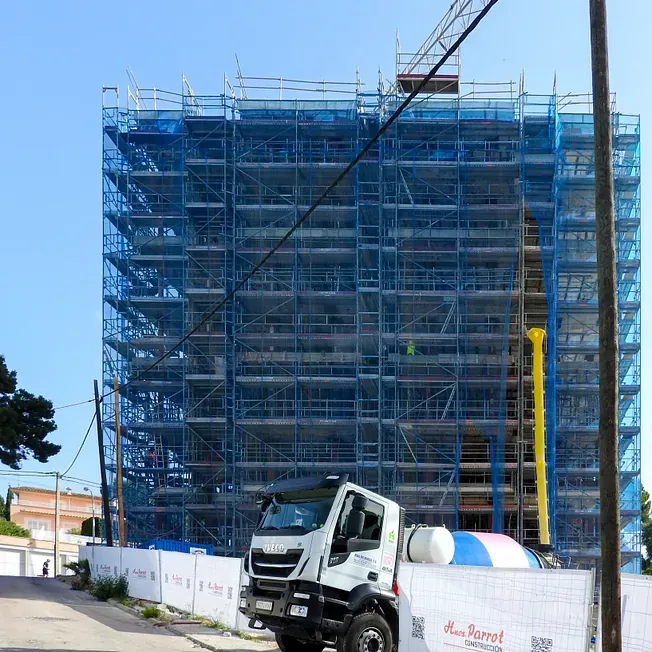 Edificio en construcción, protegido por andamios y redes azules. Un camión blanco aparcado enfrente.