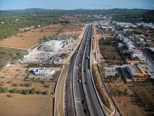 Vista aérea de una carretera que atraviesa un paisaje con escasos edificios, construcciones y colinas.
