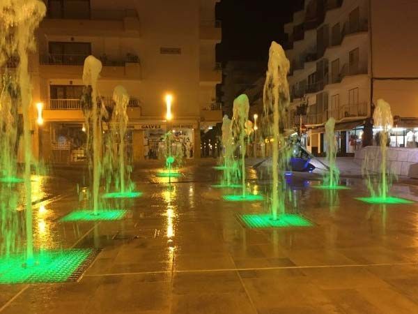 Fuentes con luces verdes que arrojan agua en una plaza de la ciudad por la noche.