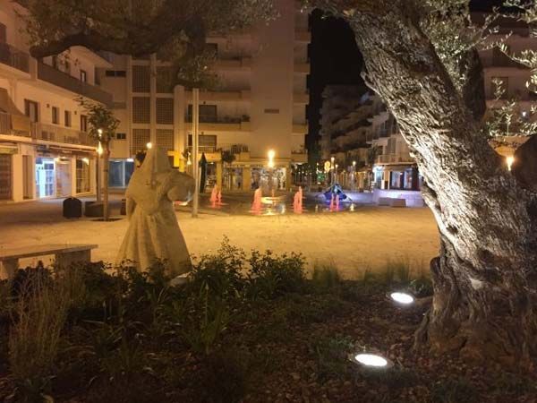 Plaza nocturna con estatua, árbol iluminado, bancos y edificios.