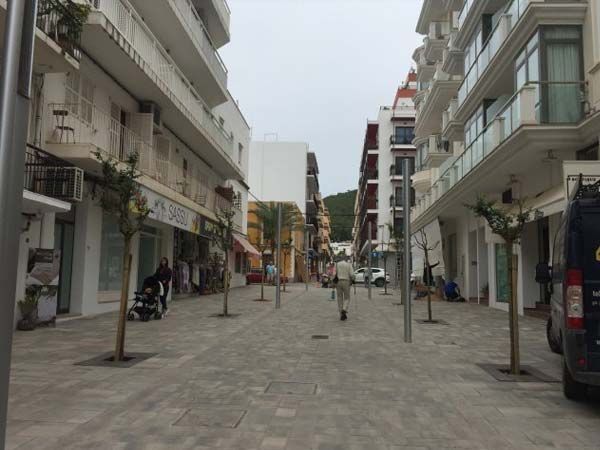 Calle bordeada de edificios blancos, tiendas y pequeños árboles, con peatones y coches a lo lejos en un día nublado.
