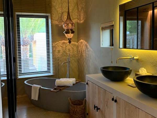 Baño con bañera gris, dos lavabos negros, tocador de madera y lámpara colgante ornamentada.