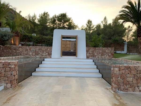 Escalones de piedra que conducen a un arco blanco, flanqueado por muros de piedra. Árboles y una colina al fondo.