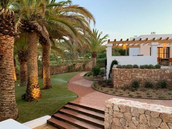 Palmeras enmarcan un sendero de piedra que conduce a un edificio blanco con pérgola. Césped verde y muro de piedra.