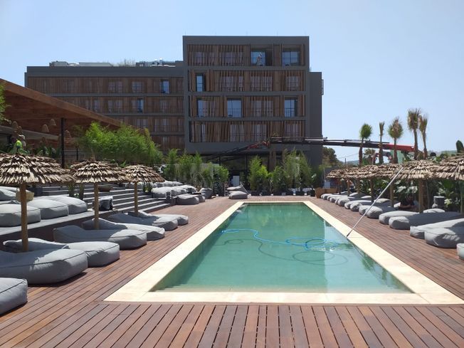 Piscina del hotel con tumbonas, sombrillas de paja y un edificio moderno al fondo.