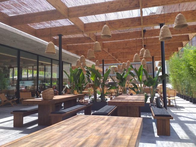 Comedor al aire libre con mesas y bancos de madera, bajo un techo de listones de bambú con lámparas colgantes y vegetación.