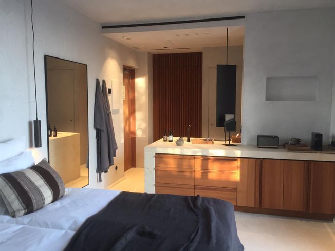 Dormitorio con paredes blancas, armarios de madera, cama y entrada al baño. La luz del sol realza la madera.