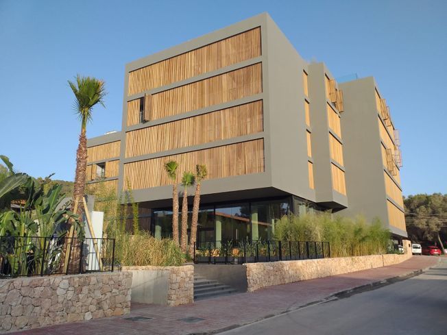Edificio gris moderno con detalles de bambú, entrada con escalones y plantas tropicales en el exterior.