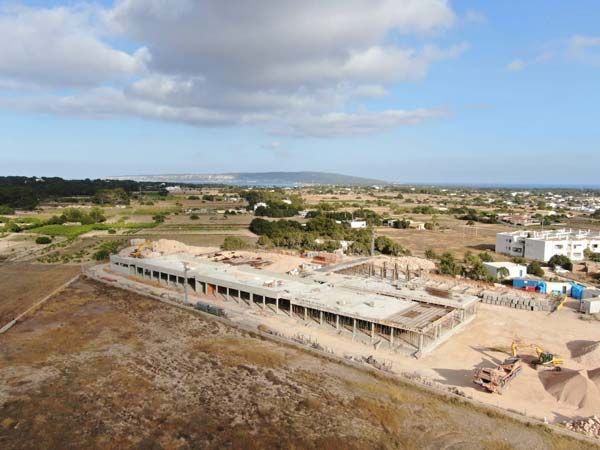Sitio de construcción con estructura de edificio, rodeado de tierra firme y edificios distantes bajo un cielo nublado.