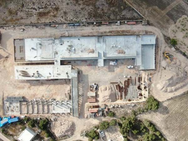 Vista aérea de un edificio en construcción, cimientos de hormigón y materiales en un terreno polvoriento.