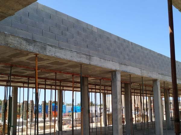 Obra en construcción con columnas de hormigón, muro de block y techo parcialmente construido, sostenido por postes metálicos.