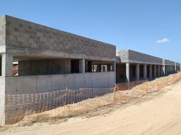 Sitio de construcción con edificio de hormigón largo y rectangular en construcción sobre un camino de tierra, cielo despejado.