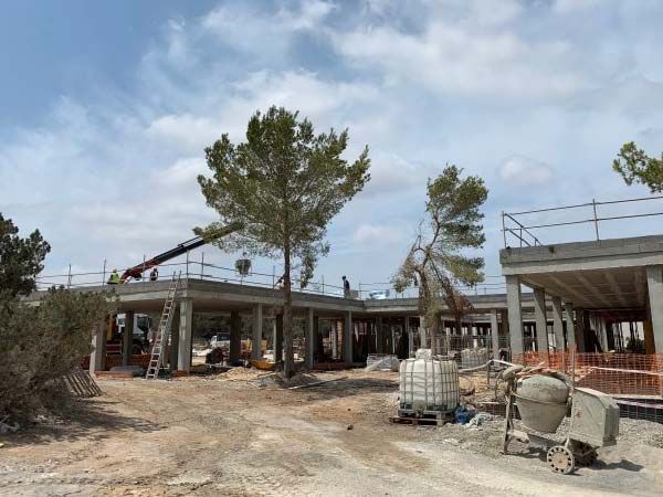 Obra con estructuras de hormigón, árboles y trabajadores. Cielo azul.