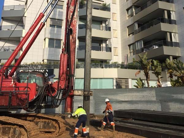 Obreros de la construcción operando maquinaria, instalando pilotes cerca de un edificio.