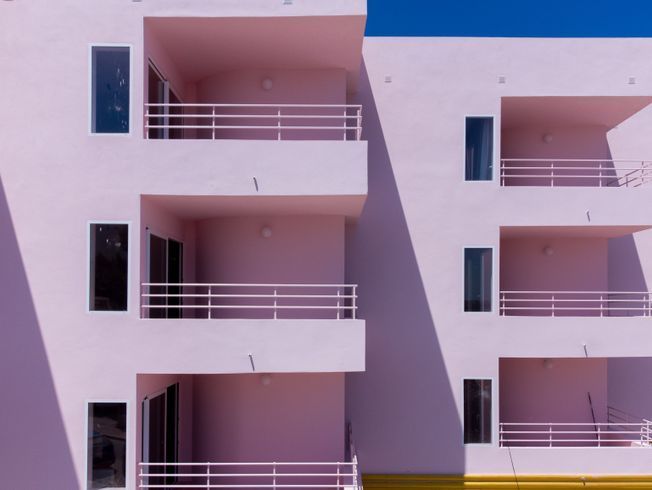 Hotel rosa con balcones, piscina y coche clásico junto al mar bajo un cielo azul.