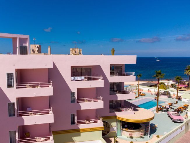 Hotel rosa con balcones, piscina y coche clásico junto al mar bajo un cielo azul.