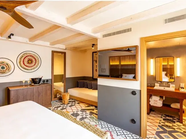 Habitación de hotel con cama, zona de estar y acceso al baño. Tonos de blanco y madera natural con arte decorativo en la pared.